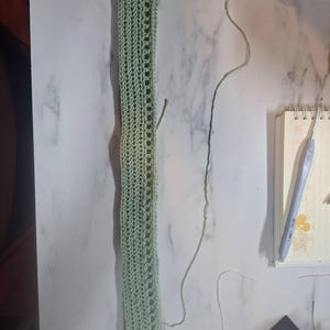 May include: A light green, rectangular crochet piece with a series of openwork rectangles down the center. The crochet work is lying on a white marble surface. A crochet hook and notebook are visible in the background.