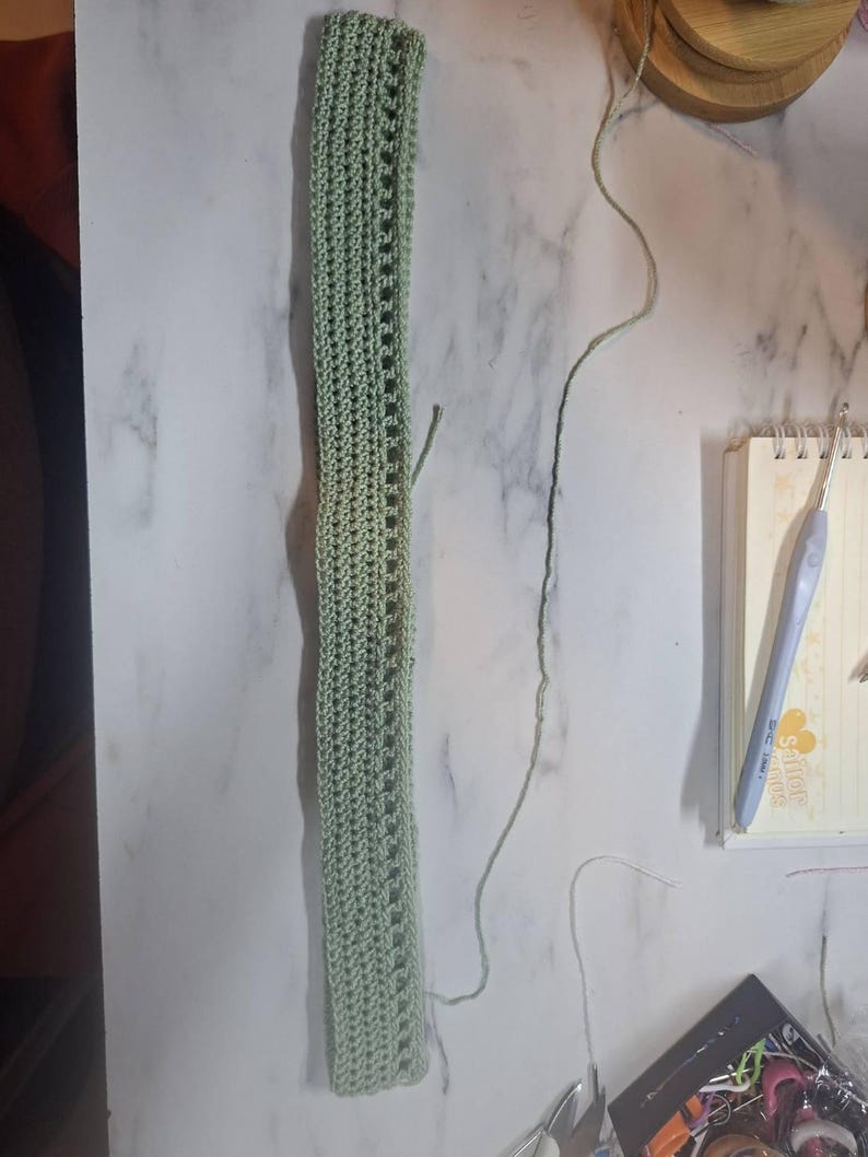 May include: A light green, rectangular crochet piece with a series of openwork rectangles down the center. The crochet work is lying on a white marble surface. A crochet hook and notebook are visible in the background.