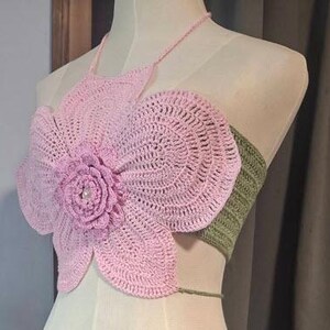 May include: A handmade crochet top in a floral design. The top features a large, light pink flower with a detailed center, and a green band at the back. The halter top is displayed on a mannequin.