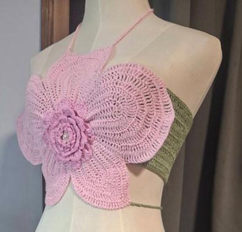 May include: A handmade crochet top in a floral design. The top features a large, light pink flower with a detailed center, and a green band at the back. The halter top is displayed on a mannequin.