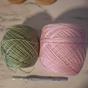 May include: Two balls of yarn, one green and one pink, sit next to a crochet hook. The yarn is tightly wound, and the hook has a gray handle. Wooden spools of white yarn are in the background.