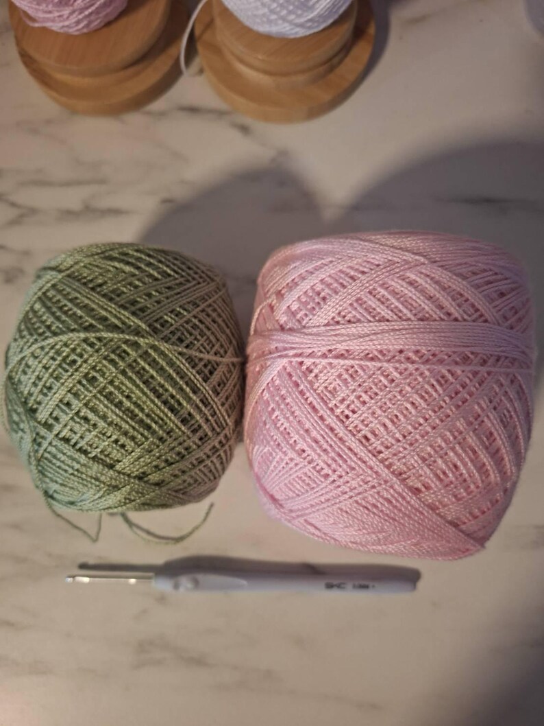 May include: Two balls of yarn, one green and one pink, sit next to a crochet hook. The yarn is tightly wound, and the hook has a gray handle. Wooden spools of white yarn are in the background.