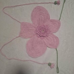 May include: A handmade, light pink crochet flower with a detailed petal design. The flower has a small, central flower with a pearl-like bead. Attached are pink strings and green stems with small tassels. The craftwork is displayed on a white surface.