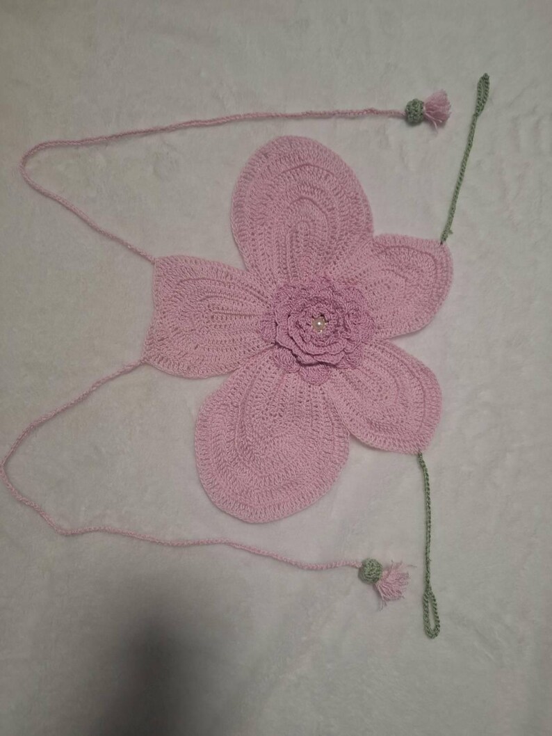 May include: A handmade, light pink crochet flower with a detailed petal design. The flower has a small, central flower with a pearl-like bead. Attached are pink strings and green stems with small tassels. The craftwork is displayed on a white surface.