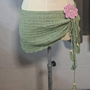 May include: A light green crochet wrap with a scalloped edge, adorned with a large pink flower and dangling tassels. The wrap is displayed on a mannequin, showcasing its intricate open weave design and decorative elements.