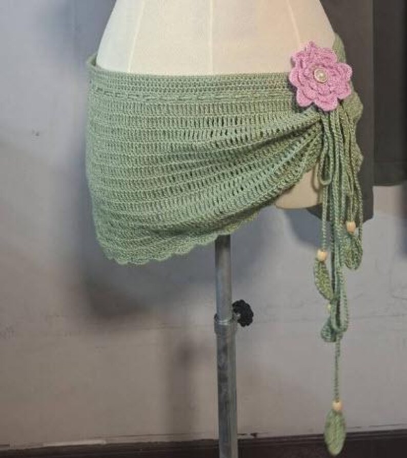 May include: A light green crochet wrap with a scalloped edge, adorned with a large pink flower and dangling tassels. The wrap is displayed on a mannequin, showcasing its intricate open weave design and decorative elements.