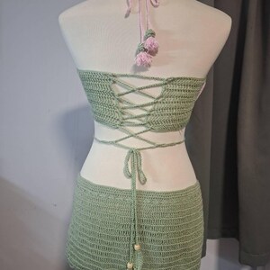 May include: A two-piece crochet set in a light green hue, displayed on a mannequin. The set includes a halter top with a criss-cross back and a matching mini skirt. Pink accents are visible on the halter straps and tassels.