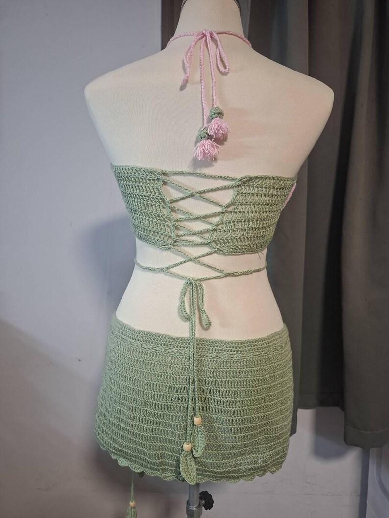 May include: A two-piece crochet set in a light green hue, displayed on a mannequin. The set includes a halter top with a criss-cross back and a matching mini skirt. Pink accents are visible on the halter straps and tassels.