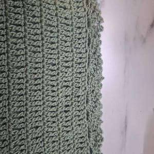 May include: Close-up of a sage green crochet fabric. The open weave pattern is visible, with vertical rows and a scalloped edge. The texture is soft and the color is a muted green.