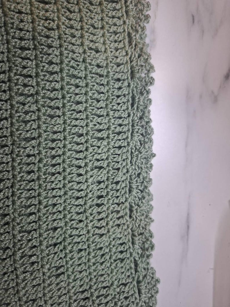 May include: Close-up of a sage green crochet fabric. The open weave pattern is visible, with vertical rows and a scalloped edge. The texture is soft and the color is a muted green.