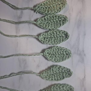 May include: Six crocheted leaf shapes in a soft green hue, each with a long, thin stem. The leaves are oval-shaped with a textured surface, likely handmade. The background is a light, marbled surface.