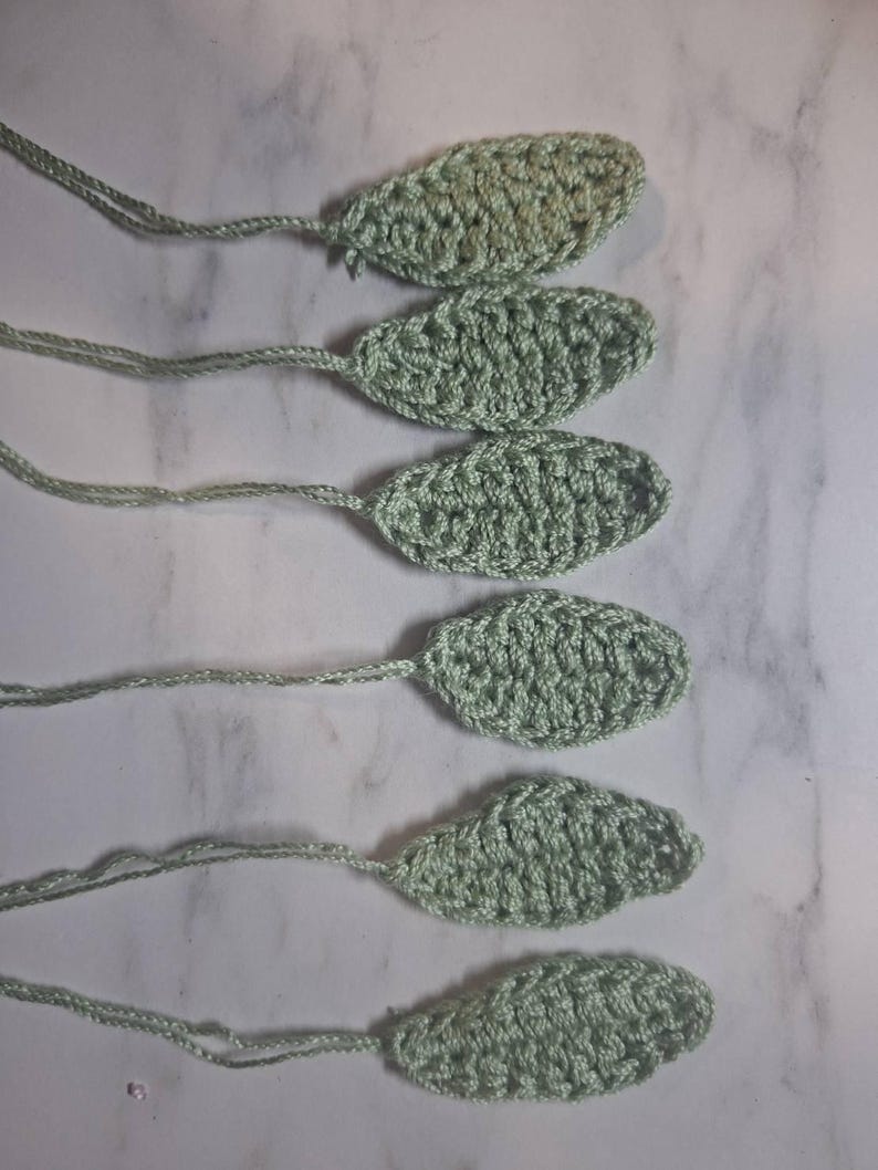 May include: Six crocheted leaf shapes in a soft green hue, each with a long, thin stem. The leaves are oval-shaped with a textured surface, likely handmade. The background is a light, marbled surface.