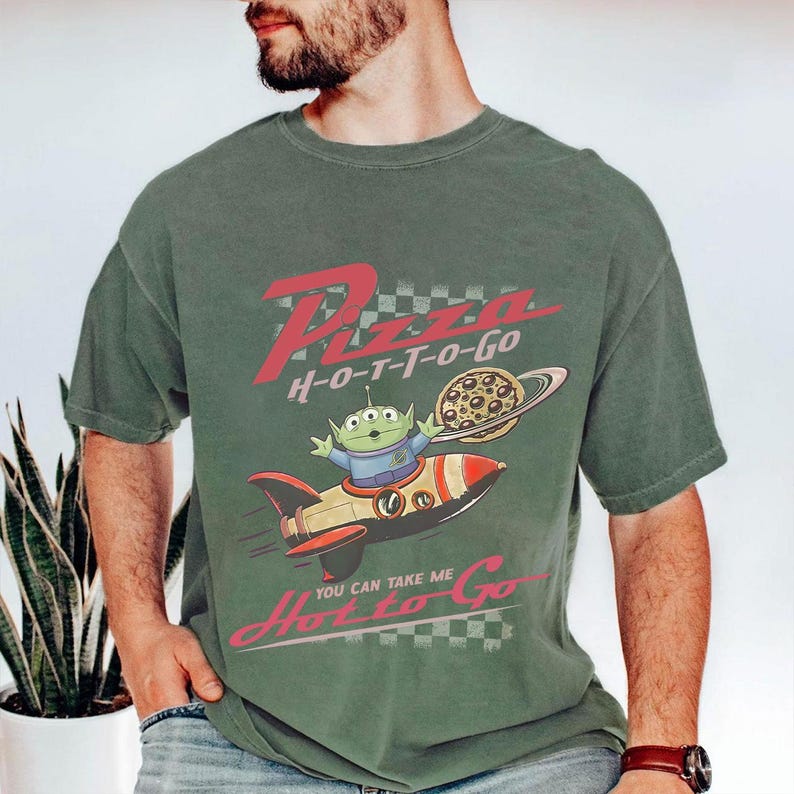 May include: Green t-shirt with a graphic featuring a cartoon alien in a rocket ship, holding a pizza. The text on the shirt reads "Pizza H-O-T-T-O-Go" and "You can take me Hot to Go".