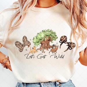 May include: Cream-colored t-shirt featuring a whimsical design with a butterfly, tree, and characters from The Lion King. The phrase "Let's Get Wild" is written in a cursive font. The butterfly and mouse ears have a leopard print.