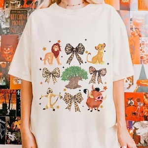 May include: Cream-colored t-shirt featuring a colorful graphic of characters and bow ties from The Lion King. The design includes Simba, Timon, Pumbaa, and a tree, with star accents.