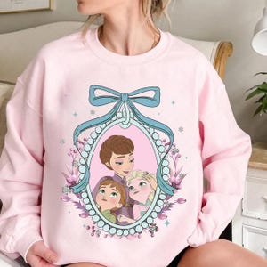 Könnte beinhalten: Hellrosa Sweatshirt mit einer gerahmten Illustration von drei Zeichentrickfiguren. Der Rahmen ist hellblau mit einer Schleife. Die Illustration zeigt eine Frau und zwei Mädchen. Das Sweatshirt hat einen Rundhalsausschnitt.