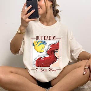 Può includere: T-shirt color crema con grafica di Ariel e Flounder de La Sirenetta, con la scritta "BUT DADDY - I Love Him" in un riquadro bordeaux. Il design presenta i capelli rossi di Ariel e i colori giallo e blu di Flounder.