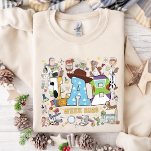 May include: A beige sweatshirt featuring a colorful graphic with the text "LAB WEEK 2026". The design includes cartoon characters and lab equipment. The letters "LAB" are in a playful, blocky style.