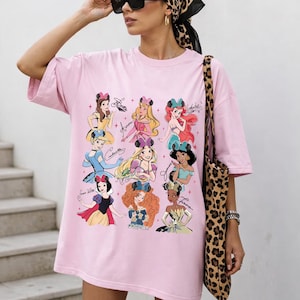 Könnte beinhalten: Hellrosa T-Shirt mit einer Grafik von neun Cartoon-Prinzessinnen und ihren Unterschriften. Die Prinzessinnen sind in verschiedenen Outfits dargestellt. Das T-Shirt ist übergroß und hat kurze Ärmel.