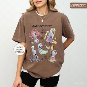 以下が含まれることがあります： エスプレッソ色のTシャツ。映画「アナと雪の女王」のオラフが様々なコスチュームを着たグラフィックが特徴です。「OLAF PRESENTS...」の文字がイラストの上にあります。左側には「MORE COLOR OPTIONS」と書かれたボタンがあります。