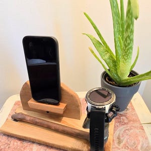 May include: A wooden phone stand with a black smartphone and a smartwatch with a black band. The stand has a natural wood finish and a small shelf for the phone. An aloe vera plant in a gray pot is also visible.