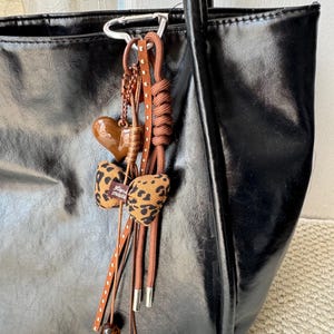Può includere: Borsa tote nera lucida, con un ciondolo decorativo. Il ciondolo include un cuore marrone, un fiocco leopardato e cordoncini marroni con perline. Il testo "Hand made" è visibile sul fiocco.