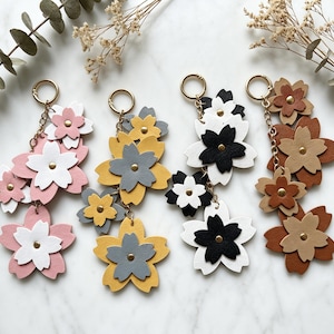 May include: Four keychains featuring layered flower shapes made of leather-like material. Each keychain has a gold-tone metal ring and chain. The flowers come in color combinations of pink and white, yellow and gray, black and white, and brown and tan.