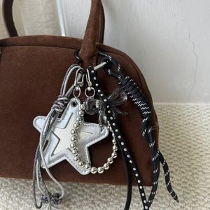 May include: A brown suede handbag with a rounded shape and a top handle. Attached to the handle are several charms, including a silver star, a beaded strand, and a black and white cord. The charms add a decorative element to the bag.