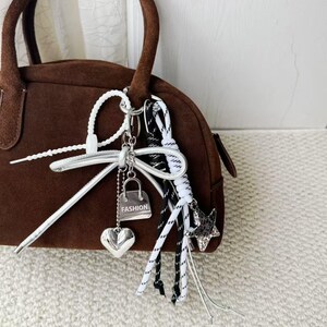 May include: A brown suede handbag with a rounded shape and a top handle. The bag is accessorized with a silver charm featuring a heart, a purse with the word "FASHION", and a star. White and black rope accents are also attached.