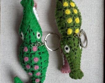 May include: Two handmade felt alligator keychains. One is green with pink spots, the other is green with yellow spots. Both have white teeth and pink accents. They are connected by a silver chain and key rings.
