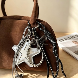 Può includere: Una borsa a mano in camoscio marrone con manico nero e vari ciondoli decorativi. I ciondoli includono una stella argentata, un fiocco trasparente e una corda bianca e nera. La borsa è su una superficie di legno.