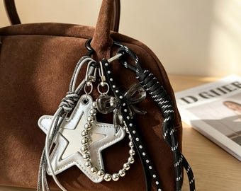 May include: A brown suede handbag with a black handle and a variety of decorative charms. Charms include a silver star, a clear bow, and a black and white rope. The bag is sitting on a wooden surface.