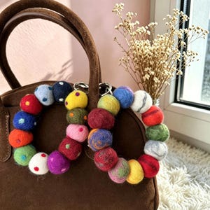 Pode incluir: Dois porta-chaves circulares feitos de bolas de lã feltrada multicoloridas em vários tons de vermelho, azul, verde, amarelo, rosa e branco. As bolas apresentam cores sólidas, bolinhas e padrões marmoreados, unidos num anel de metal prateado.