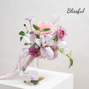May include: A wedding bouquet with pink, white, and purple flowers, including roses, carnations, and calla lilies. The bouquet is in a clear glass vase and has a matching boutonniere. The word "Blissful" is written in cursive on the image.