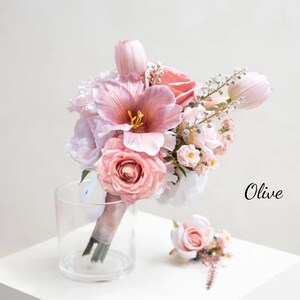 May include: A wedding bouquet with pink roses, tulips, and other flowers, plus a matching buttonhole. The bouquet is tied with a pink ribbon and placed in a clear glass vase. The word "Olive" is written in black script.
