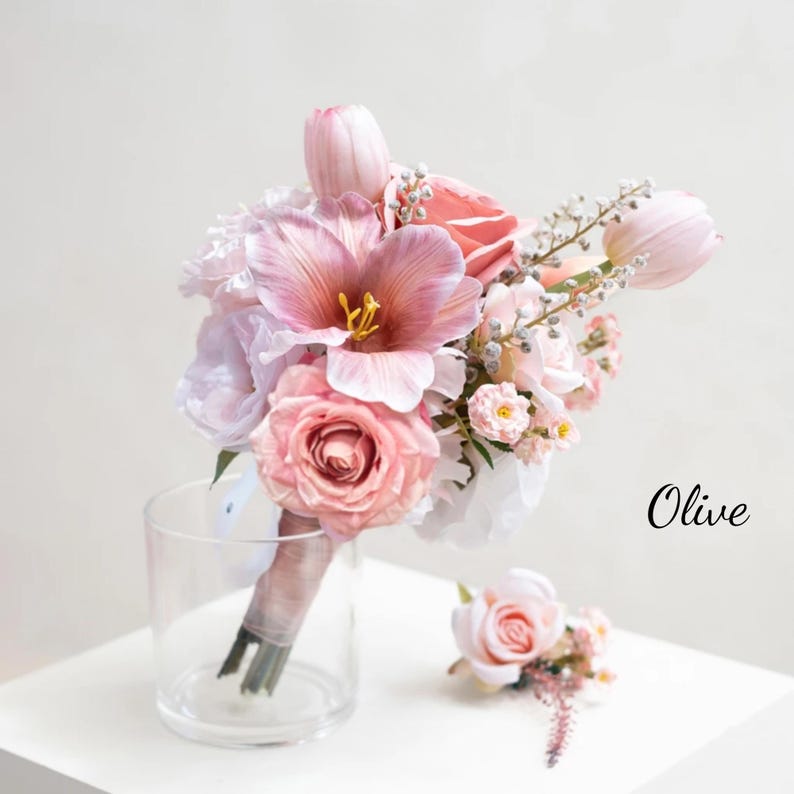 May include: A wedding bouquet with pink roses, tulips, and other flowers, plus a matching buttonhole. The bouquet is tied with a pink ribbon and placed in a clear glass vase. The word "Olive" is written in black script.