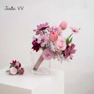May include: A floral bouquet featuring pink and purple blooms, such as roses, tulips, and anemones, with a coordinating buttonhole. The bouquet is presented in a clear glass vase and secured with a ribbon. The text "Jade VV" is visible.