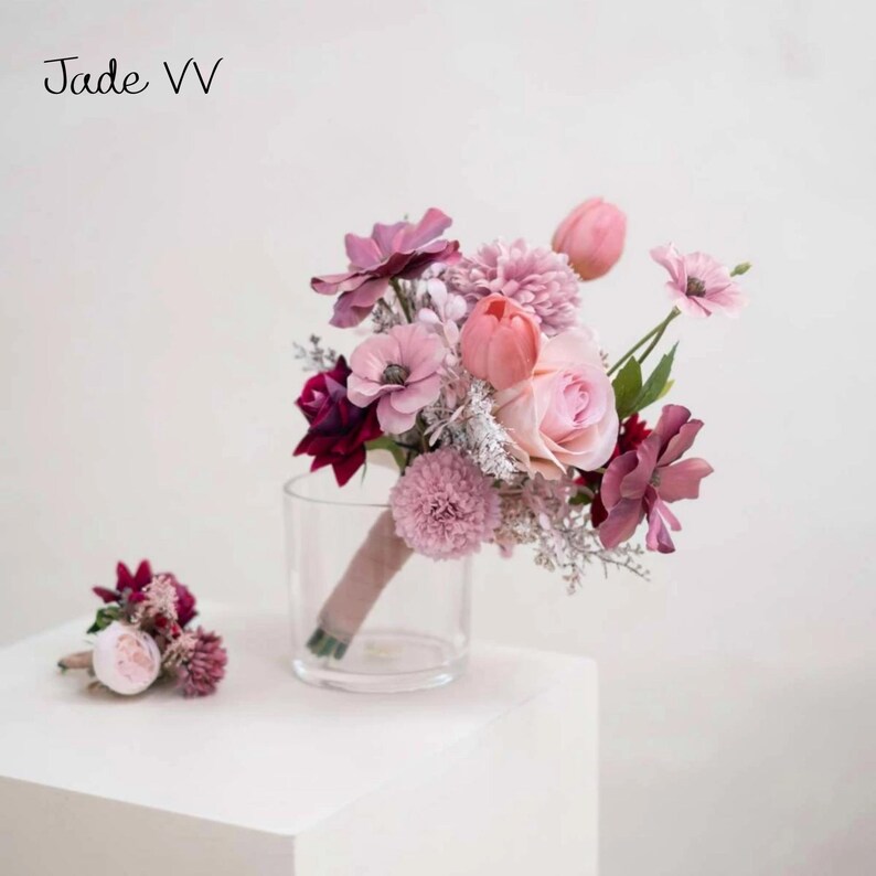 May include: A floral bouquet featuring pink and purple blooms, such as roses, tulips, and anemones, with a coordinating buttonhole. The bouquet is presented in a clear glass vase and secured with a ribbon. The text "Jade VV" is visible.