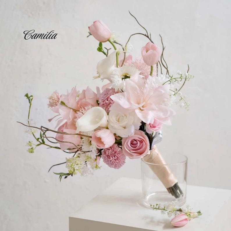 May include: A wedding bouquet of pink and white flowers, including roses, tulips, and lilies, with a ribbon-bound handle. The bouquet is in a clear glass vase. The word "Camilia" is in the upper left corner. The bouquet is a mix of flowers.