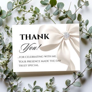 May include: A white card with the words "THANK You!" in black script and a cream-colored bow with a rhinestone center. The card also includes the text "...FOR CELEBRATING WITH ME. YOUR PRESENCE MADE THE DAY TRULY SPECIAL."