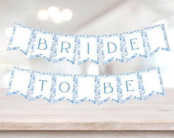 May include: A white banner with blue floral trim spelling "BRIDE TO BE". The banner is made of individual pennants, each with a blue letter. The background is blurred, with a wooden surface in the foreground.