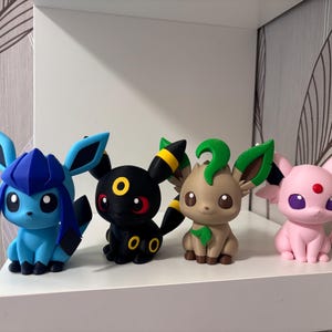Può includere: Quattro figurine colorate di Pokémon sono esposte su una mensola bianca. Le figurine includono un Glaceon blu e nero, un Umbreon nero, un Leafeon marrone e un Espeon rosa. Ogni figura ha caratteristiche e colori unici.