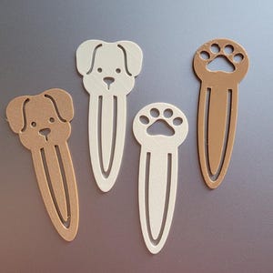 Può includere: Quattro segnalibri decorativi nei toni del marrone e del bianco. Sono visibili due segnalibri a forma di cane e due segnalibri con impronte di zampe. I segnalibri sono realizzati in materiale piatto e opaco.