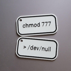 Op de afbeelding: Twee witte rechthoekige labels met afgeronde hoeken en zwarte randen. Het bovenste label leest "chmod 777" in zwarte tekst. Het onderste label leest "> /dev/null" in zwarte tekst. Elk label heeft een klein gaatje aan de bovenkant.