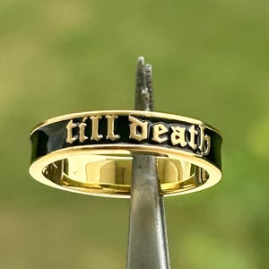 May include: A gold-colored ring with the words "till death" in black lettering. The ring has a black enamel band. The ring is held by a silver tool against a blurred green background.