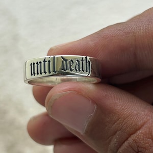 May include: A silver ring with the words "until death" engraved in a gothic font. The ring is rectangular and has a polished finish. The ring is held by a person.