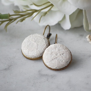 May include: A pair of round, white lace earrings with gold-colored metal frames and ear wires. The earrings are set against a soft, blurred background with white flowers and a lace doily, suggesting a delicate, feminine aesthetic.