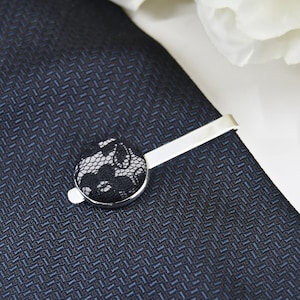 May include: A silver tie clip with a black lace button. The tie clip is on a dark blue patterned tie.