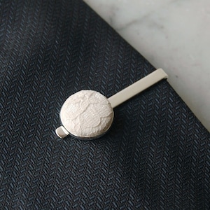 May include: A silver tie clip with a white lace button. The button is round and has a delicate lace pattern.