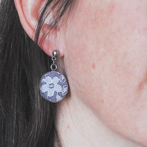 May include: A silver-toned earring with a round, button-like design. The earring features a white floral lace pattern set against a dark blue background. The earring is attached to a silver post.